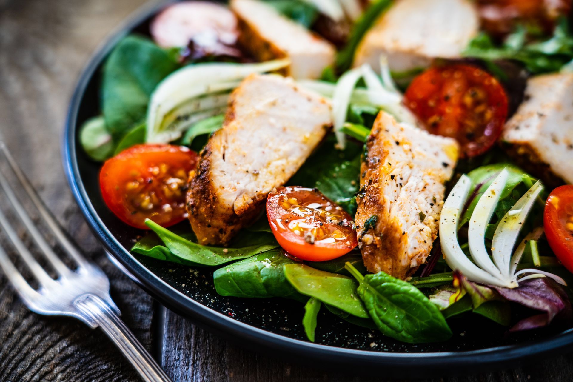 Chicken salad with tomatoes, spinach, and onion on a dark plate, near a fork.