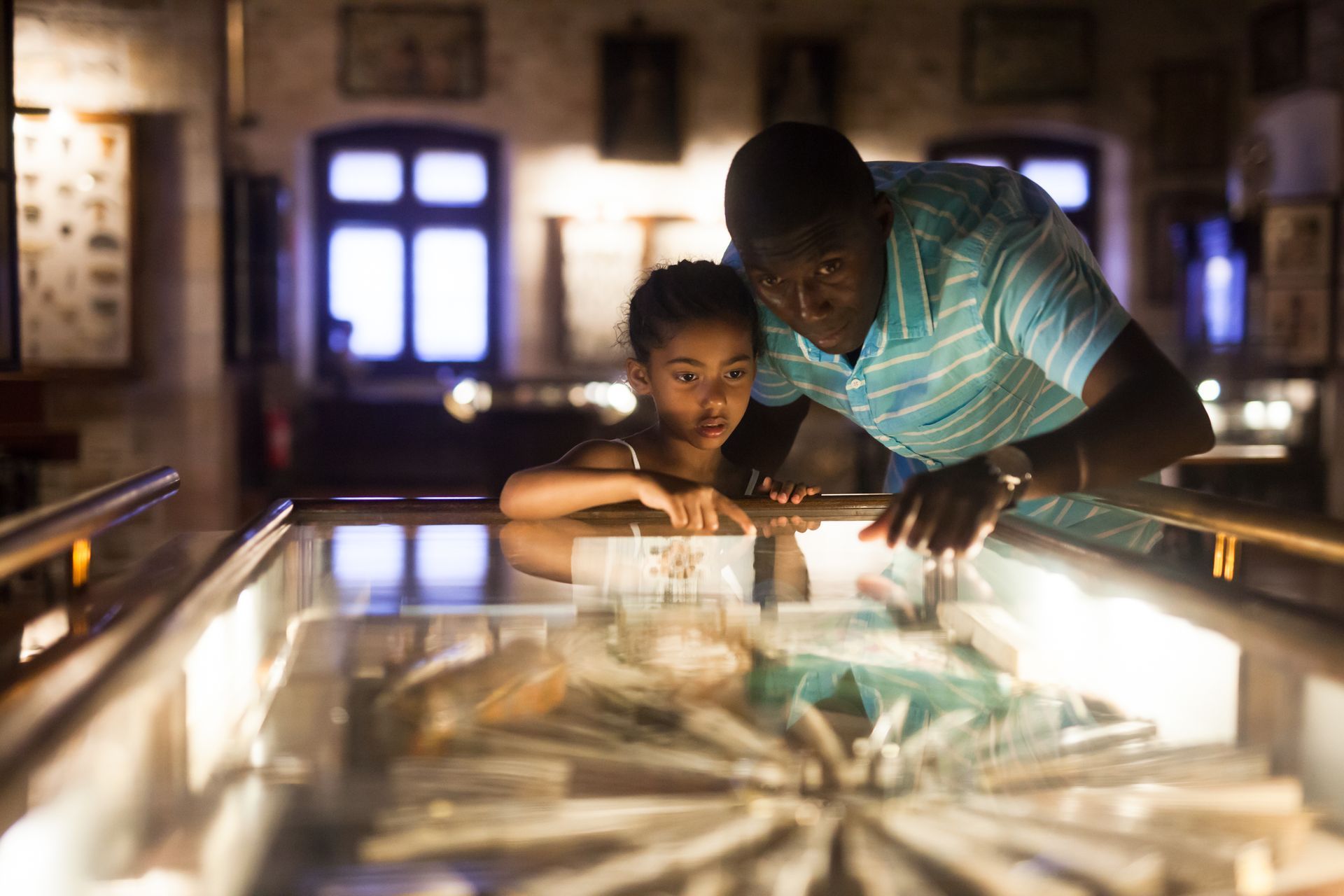 Father and daughter look at artifacts in a museum display case. They appear curious. Low lighting.