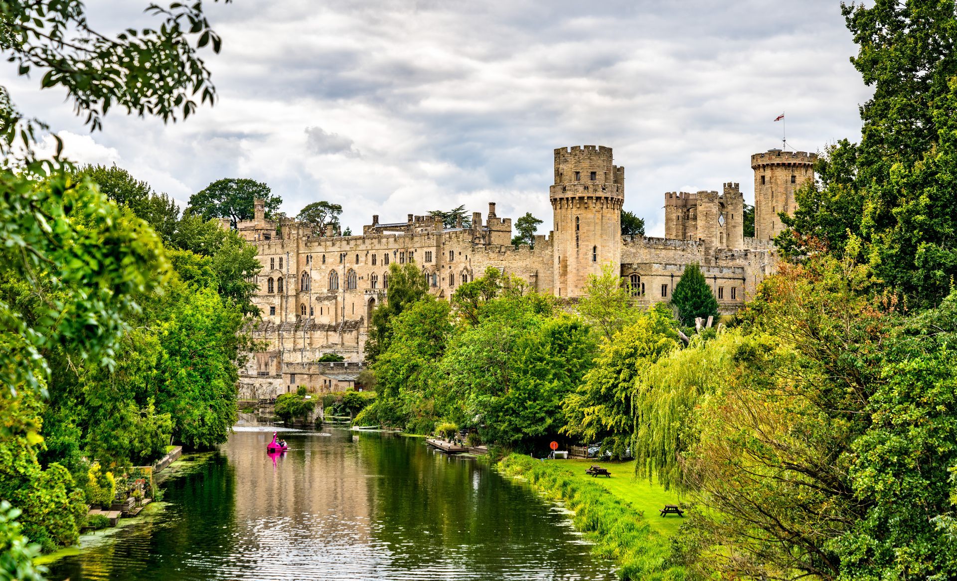 Warwick Castle, England, seen from across a river, surrounded by lush green trees under a cloudy sky.