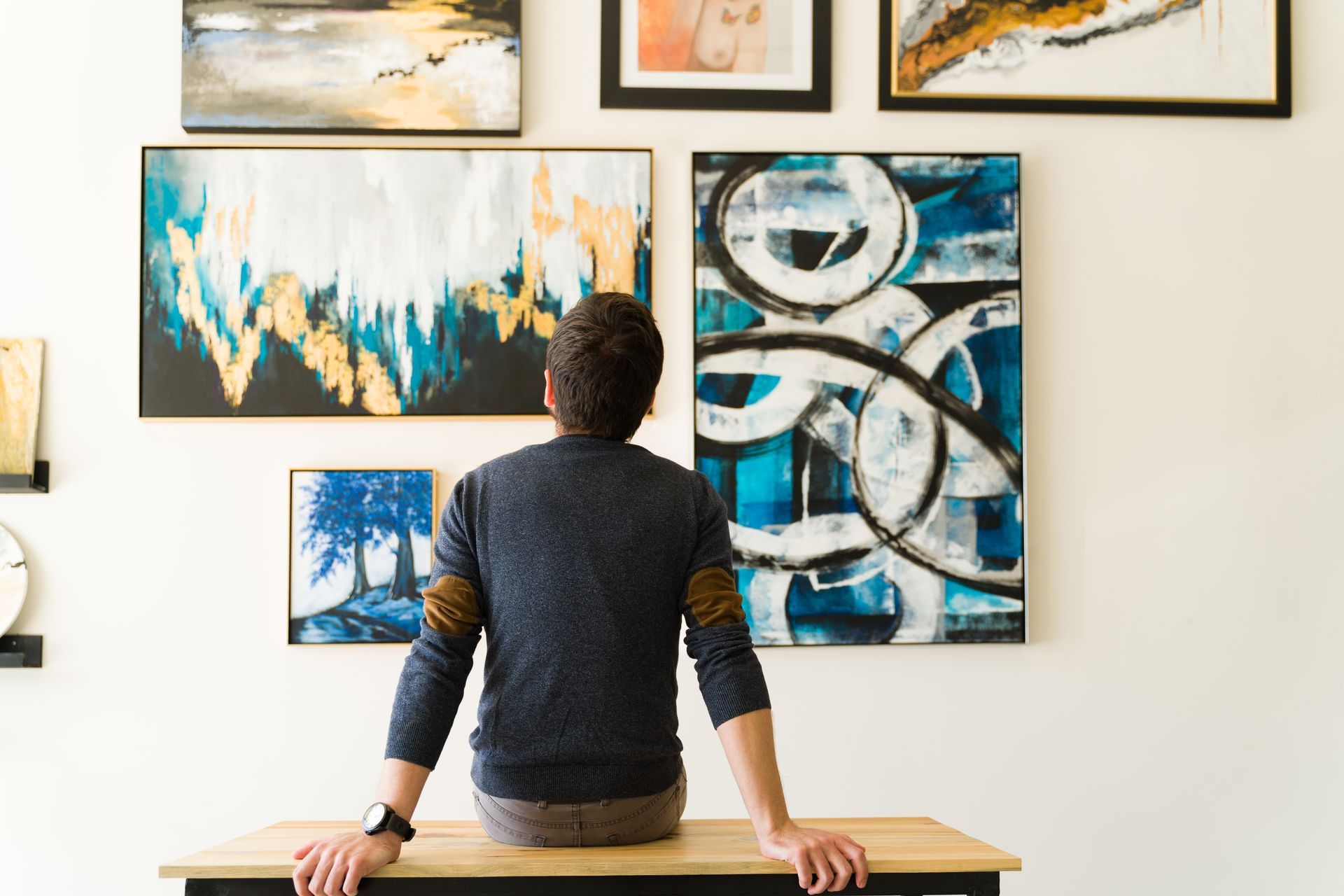 Man seated, looking at abstract art on a white wall; art features blue, gold, and white.