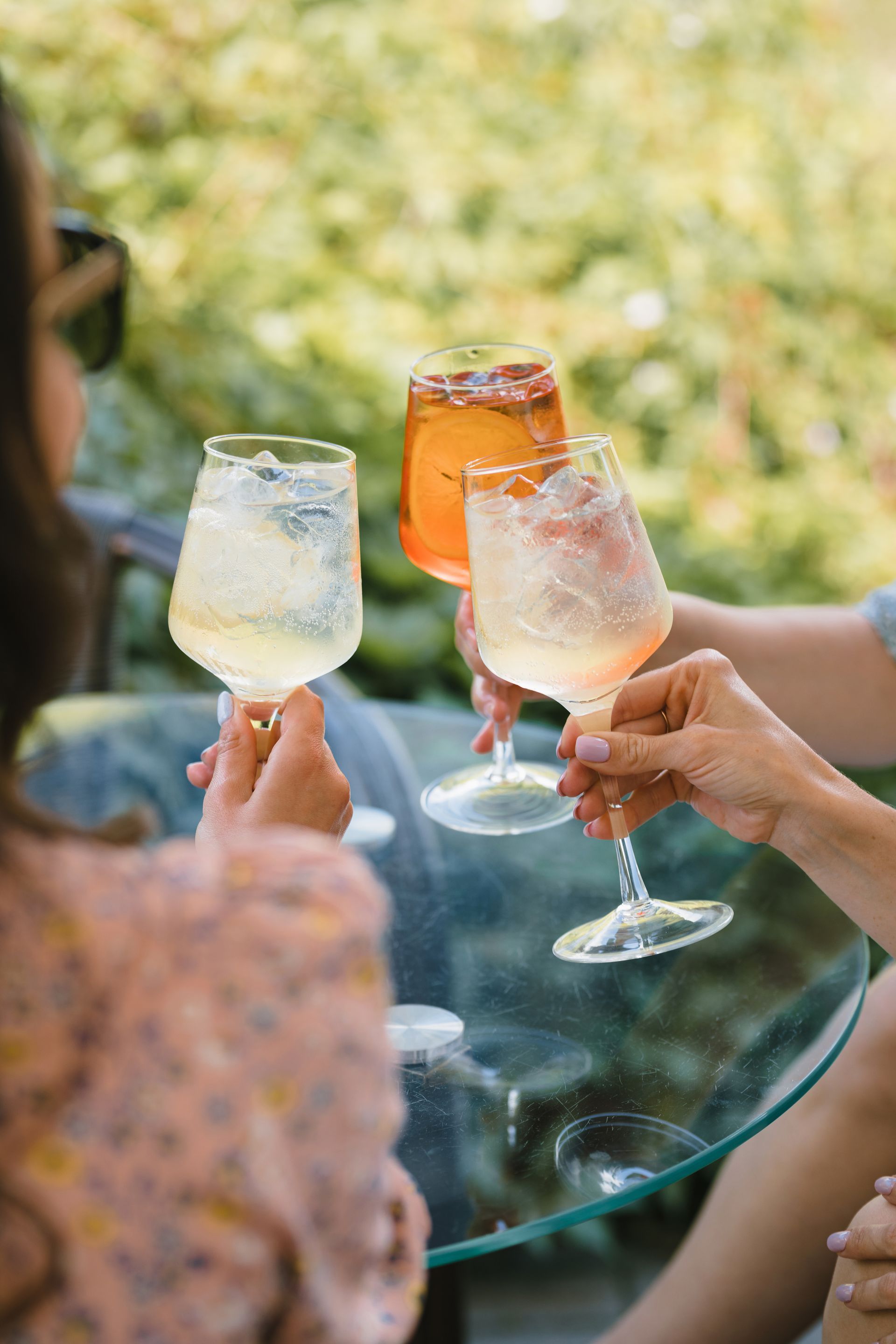 Three people clinking glasses filled with cocktails on a glass table, outdoors.