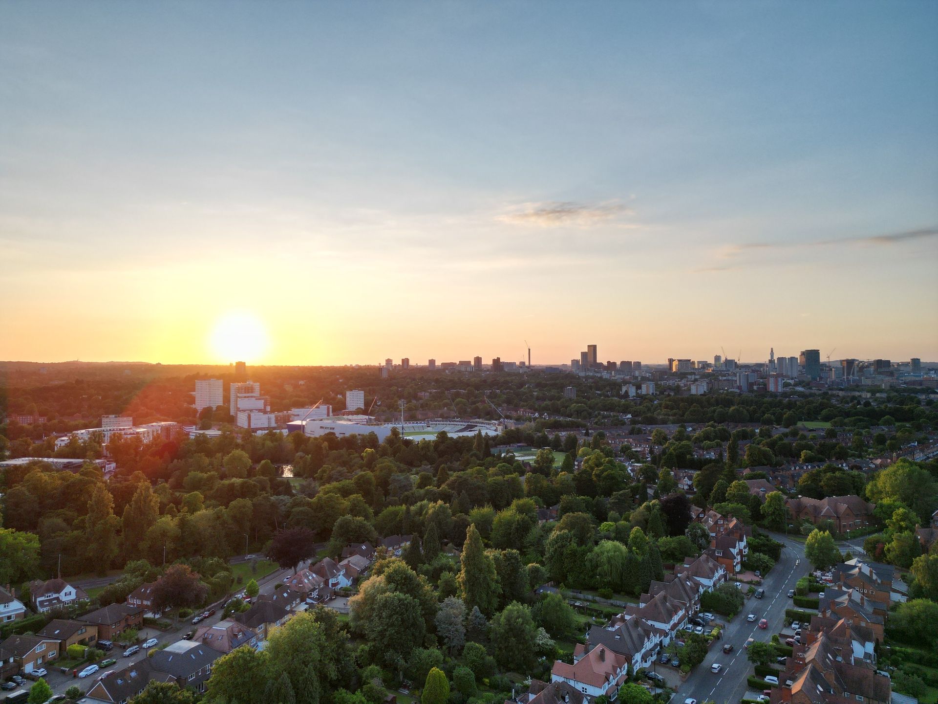 Sunset over a city skyline, with green trees and suburban homes in the foreground.