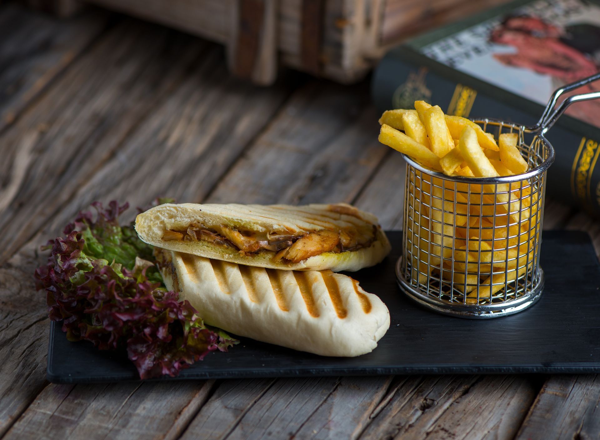 Grilled sandwich with fries in a metal basket on a black plate, set on a wooden surface with a book in the background.
