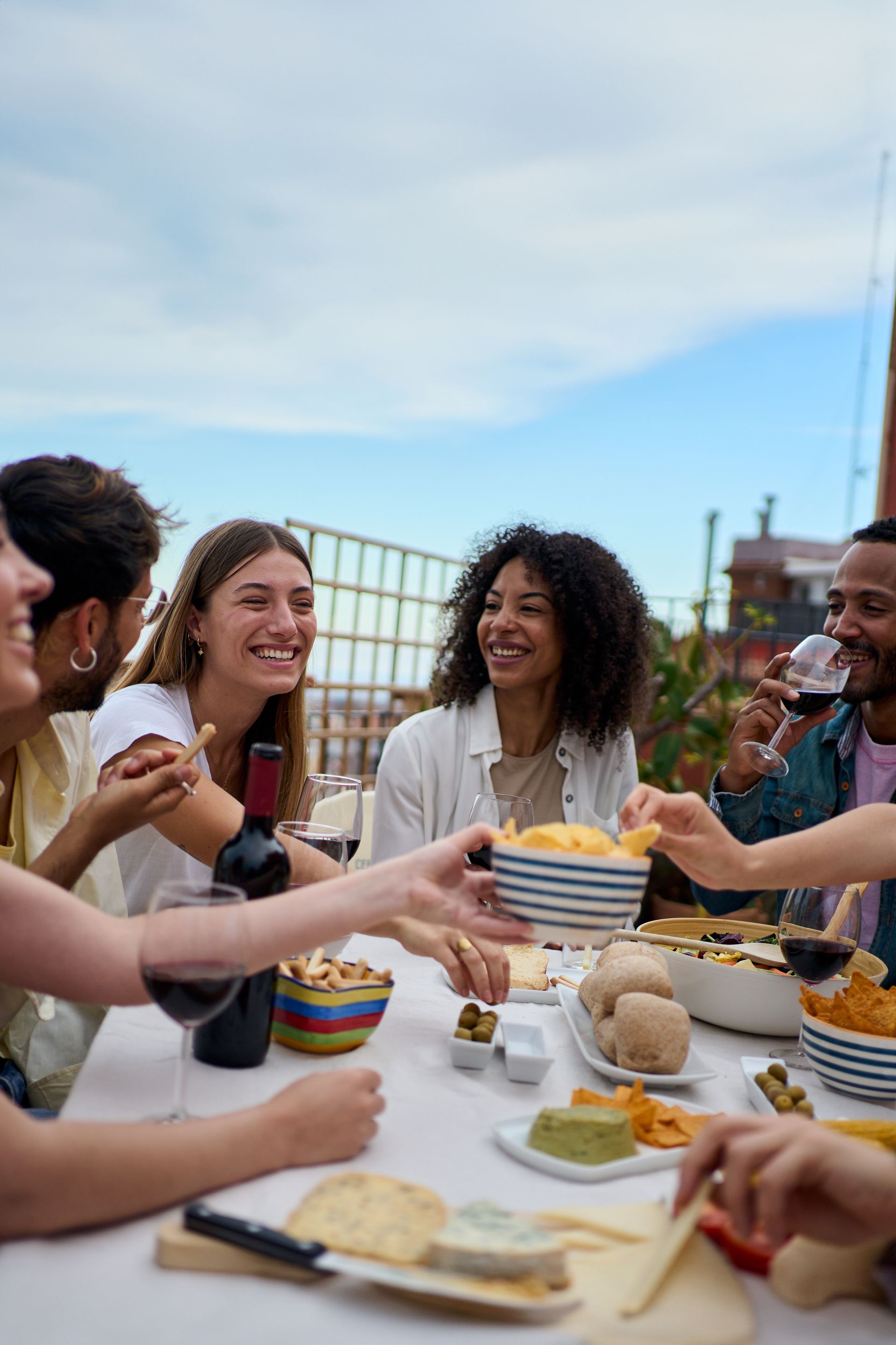 Friends sharing food and drinks on a rooftop. Smiling faces, snacks, wine, and blue sky.