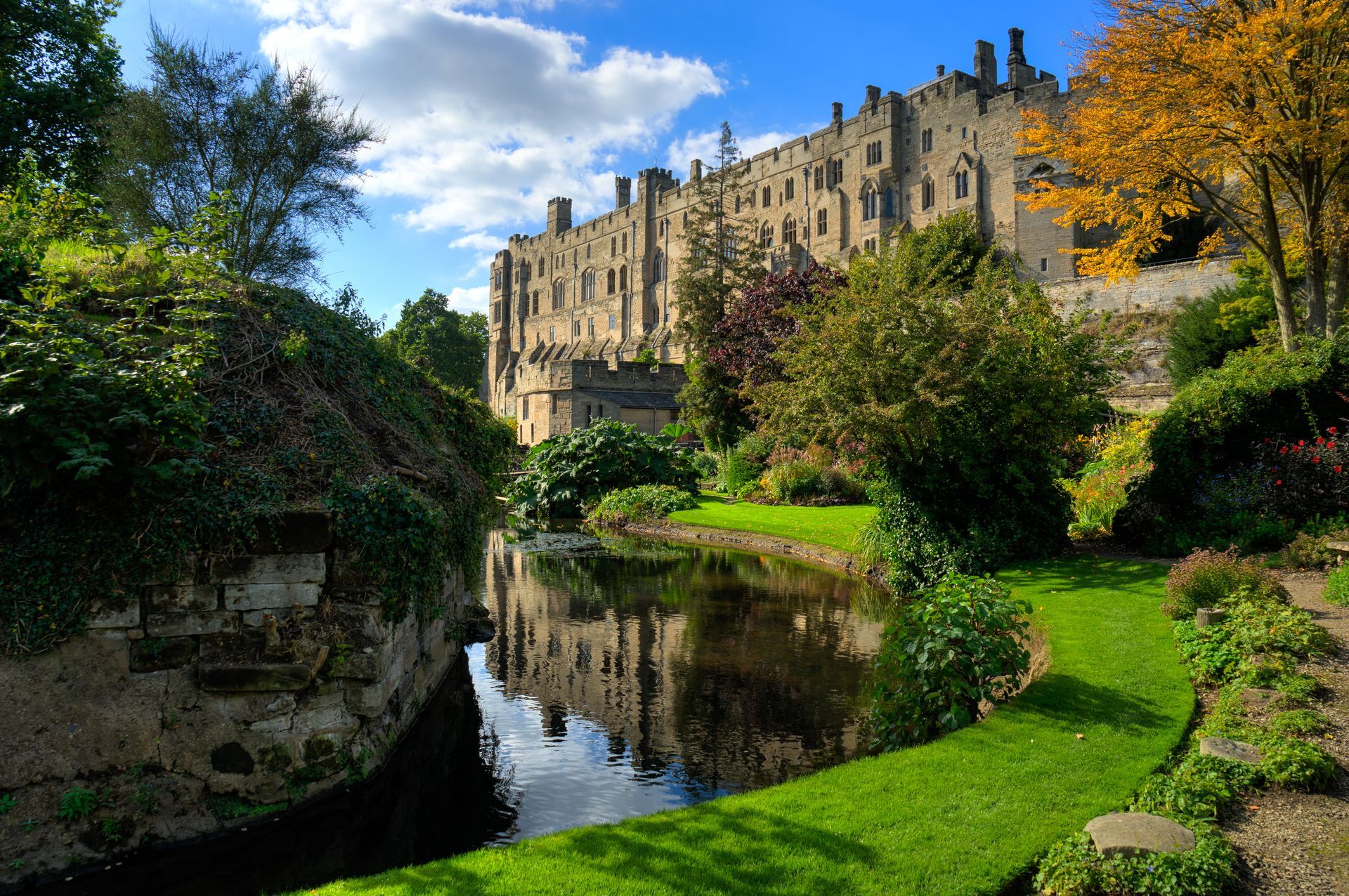 Warwick Castle reflected in a calm moat, surrounded by green grass, trees, and a blue sky with clouds.