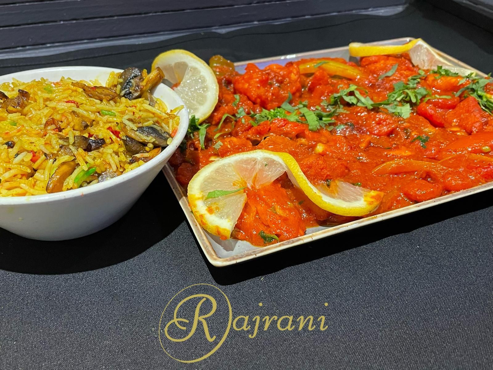 A side dish of yellow mushroom rice next to a platter of red saucy chicken garnished with cilantro and lemon slices.