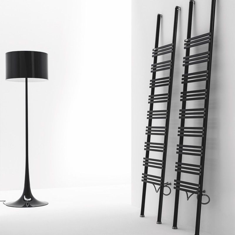 Una lampada da terra alta e nera si trova accanto a due sottili termosifoni neri a forma di scala, appoggiati a una parete bianca.
