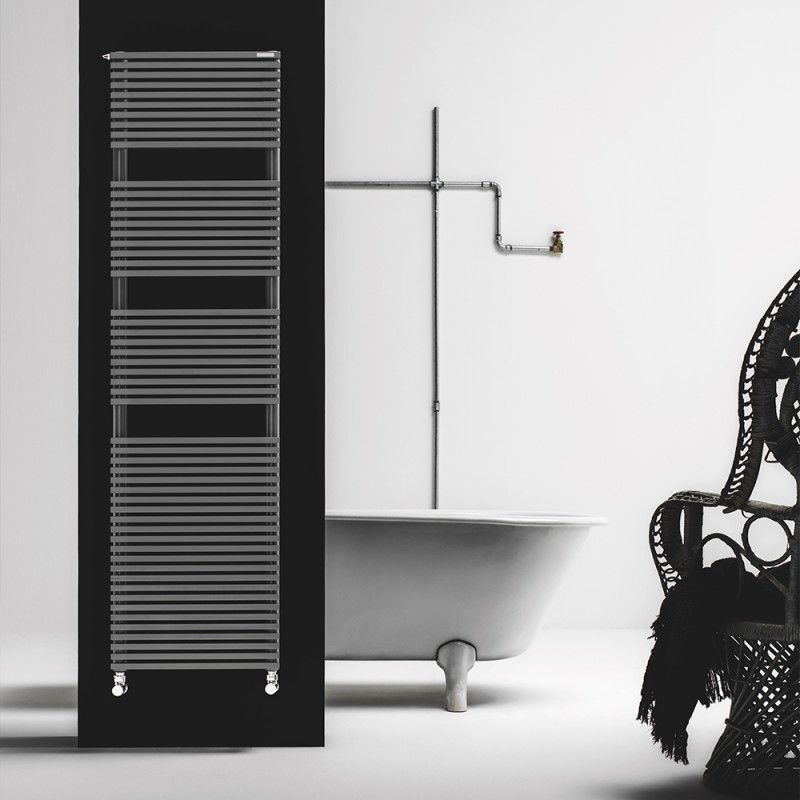 In una stanza minimalista, un alto termoarredo argentato con listelli è posizionato su un pannello nero accanto a una vasca da bagno vintage bianca con piedini a zampa di leone.