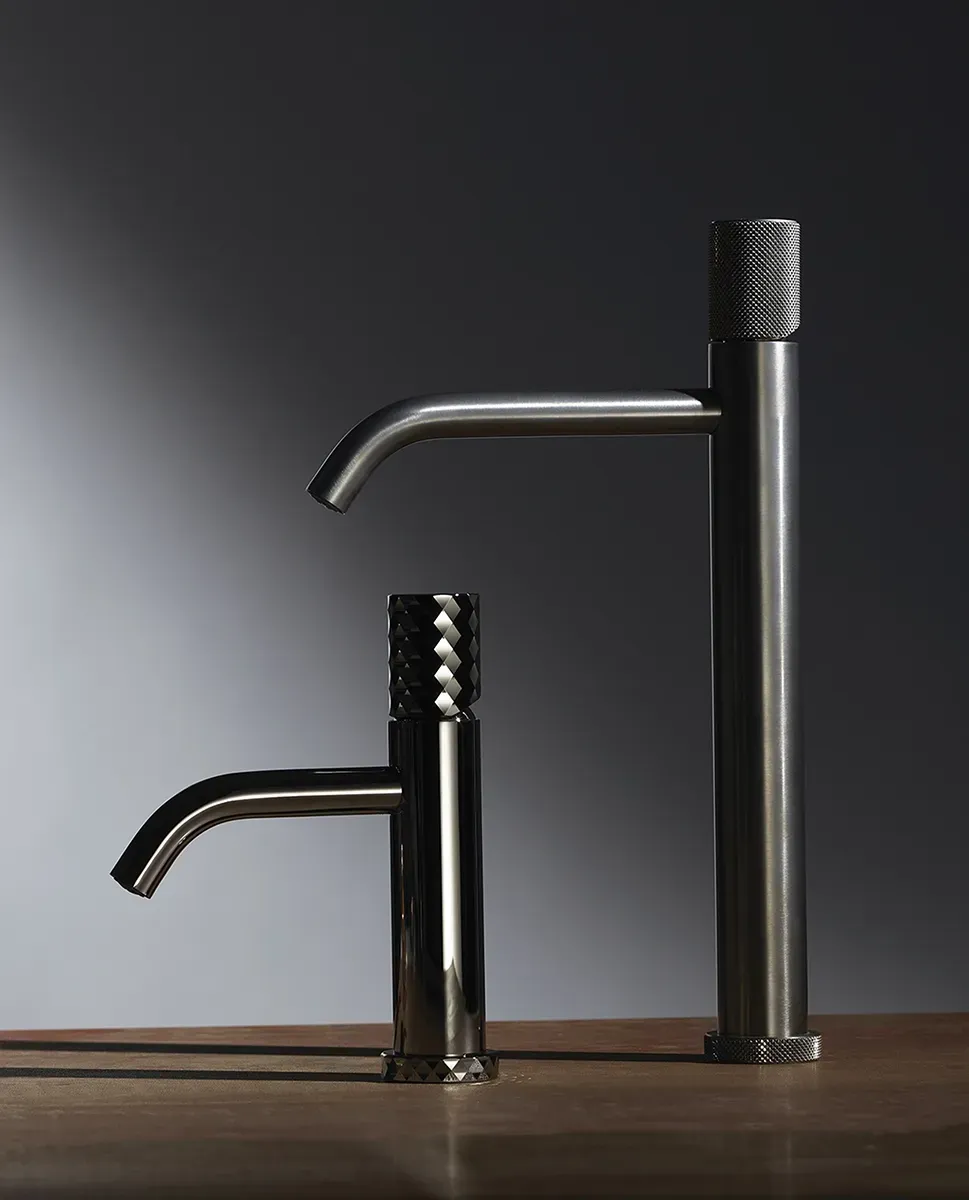 Due rubinetti moderni in metallo nichelato spazzolato con maniglie cilindriche dalla superficie zigrinata sono posizionati su una superficie in legno su uno sfondo scuro.