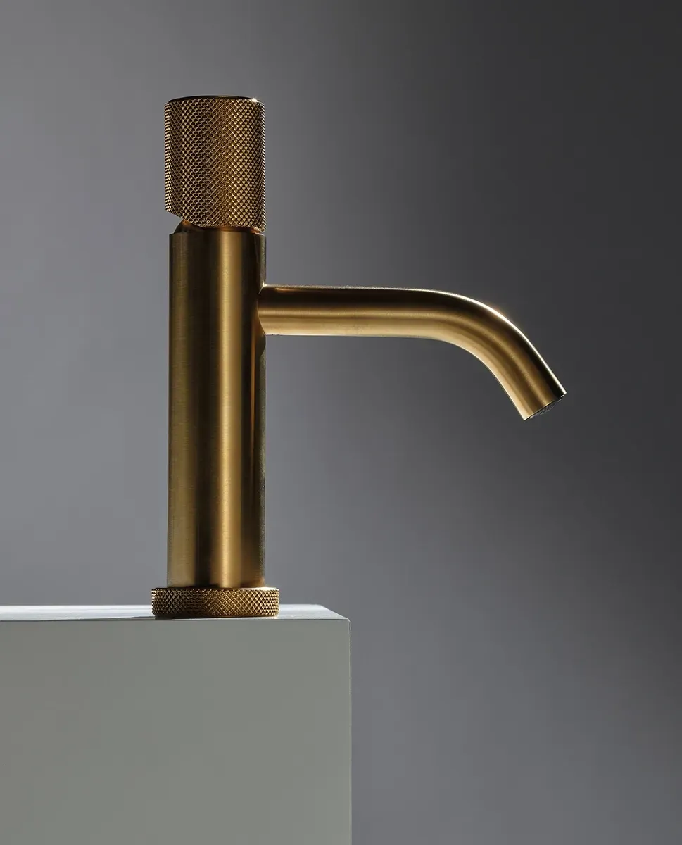 Rubinetto da bagno in oro spazzolato con maniglia e base cilindriche zigrinate, montato su una superficie bianca a contrasto con una parete grigia.
