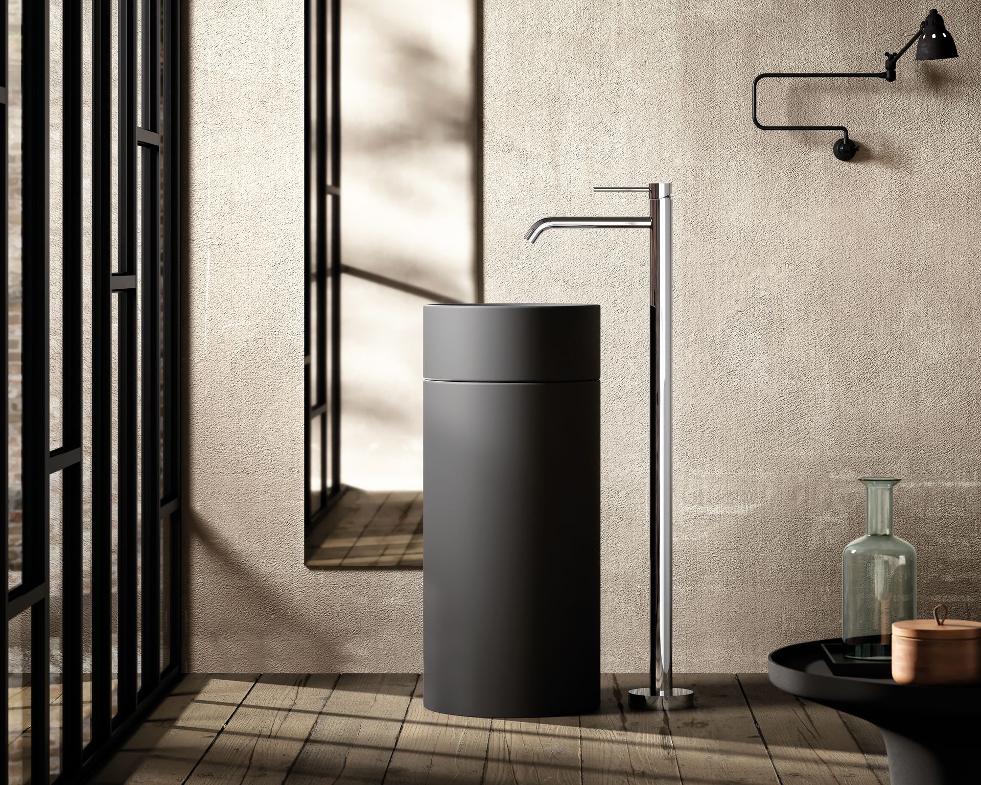 In un bagno moderno e dalle superfici materiche, un lavabo a colonna minimalista color antracite si erge accanto a un rubinetto alto e cromato a pavimento.