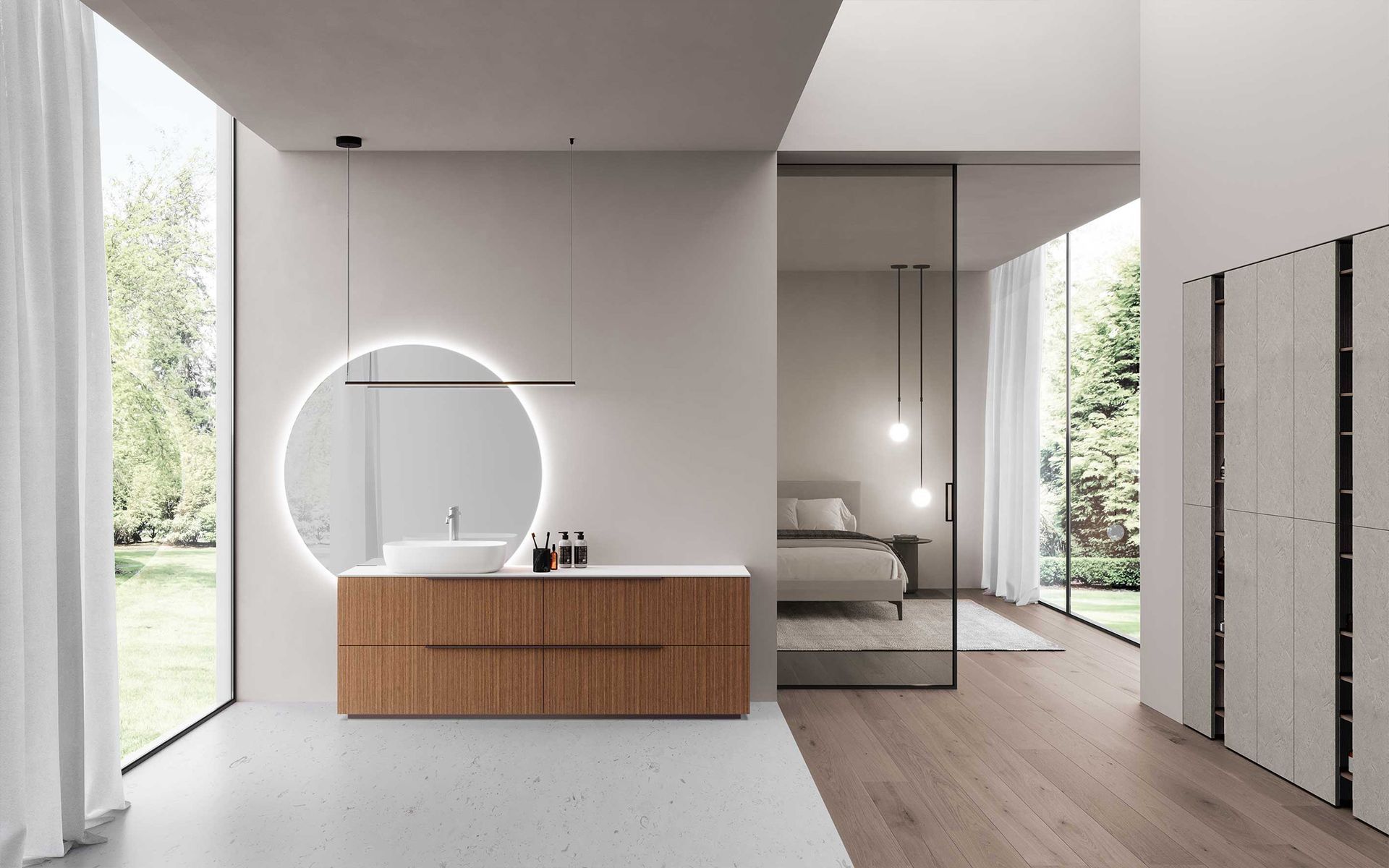 Un bagno moderno con mobile lavabo in legno e specchio rotondo luminoso, aperto su una camera da letto visibile attraverso una porta a vetri.