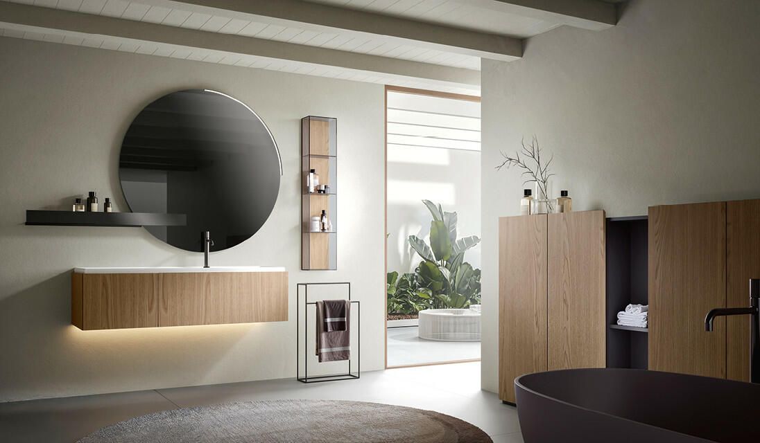 Bagno moderno con mobile lavabo sospeso in legno, specchio circolare, mobili in legno coordinati e vasca da bagno freestanding di colore scuro.