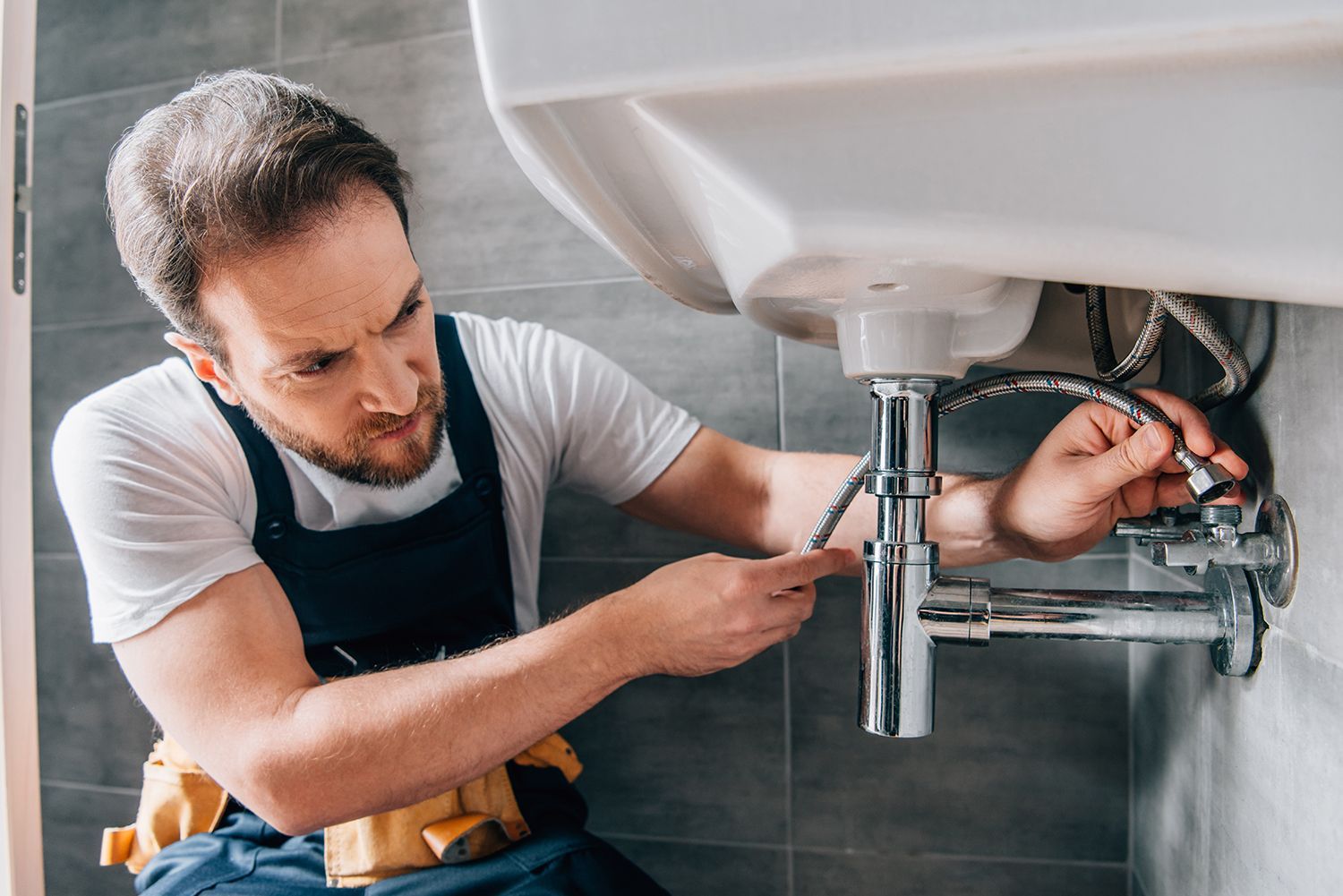 A plumber in working overalls is fixing a sink in the bathroom.