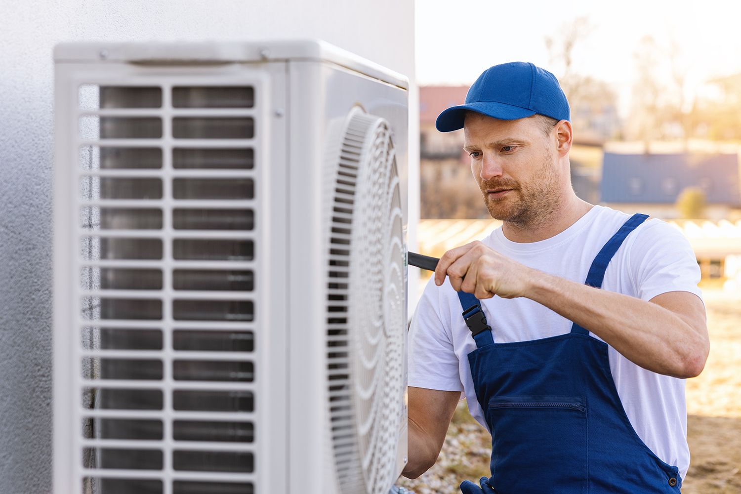 A technician is tightening outdoor AC unit components with a tool beside a house wall area