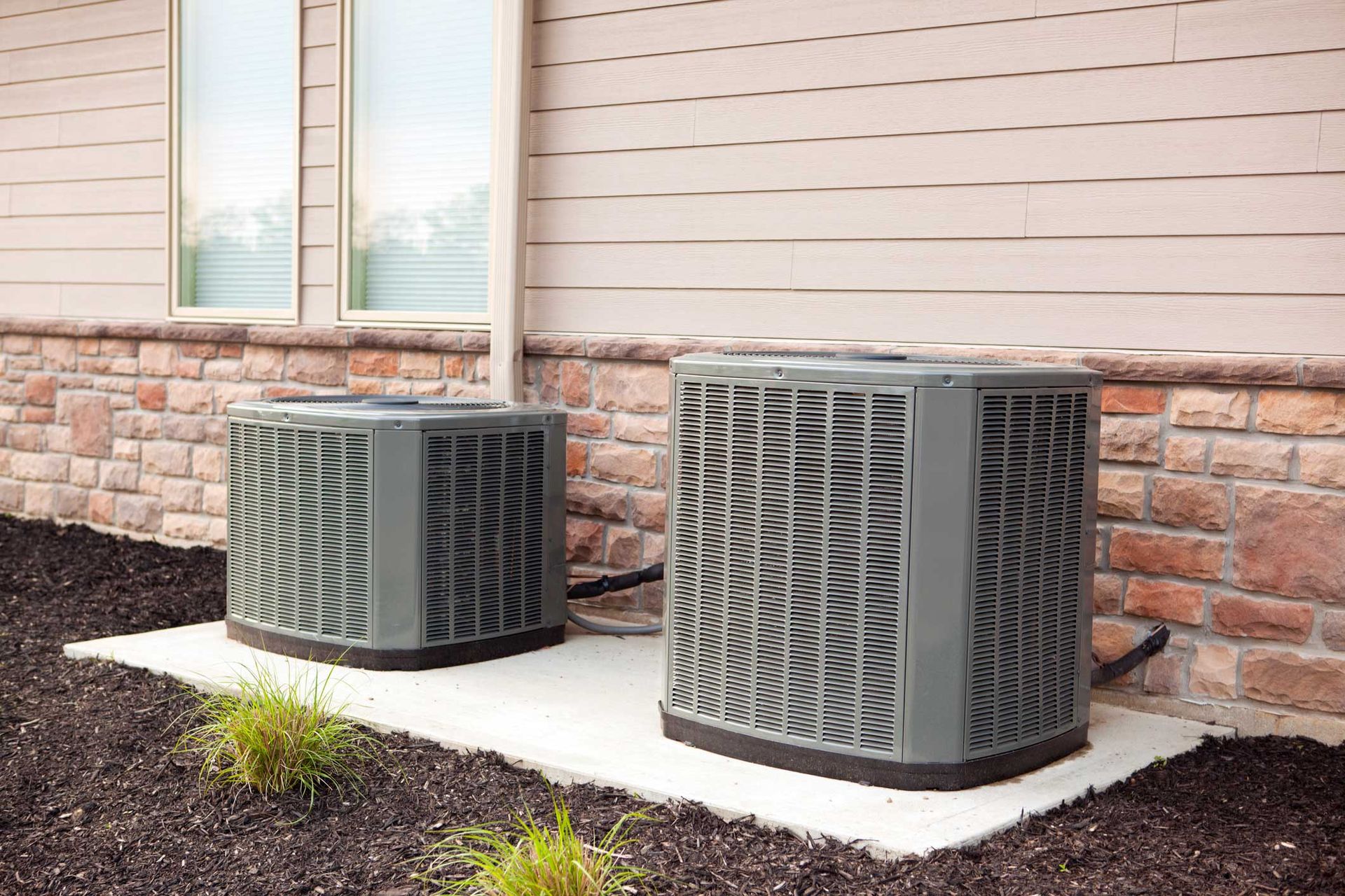 Two air conditioning units on a concrete pad next to a brick and siding wall.