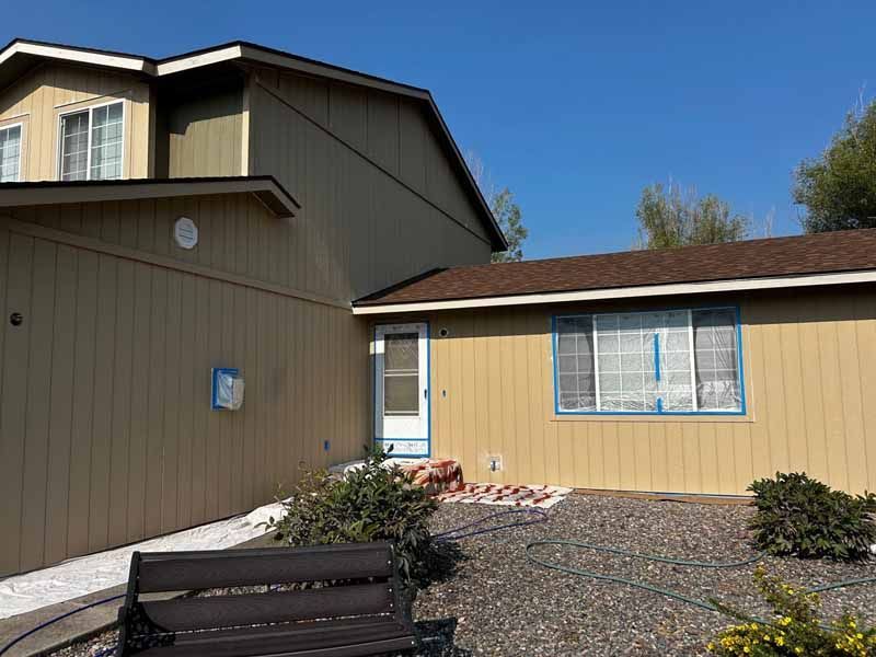 A two-story tan house with a brown roof, a gravel yard, a dark bench in the foreground, and a white screen door.