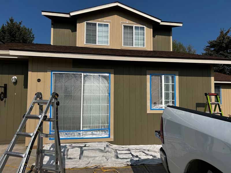 A two-story house with green siding and taped-off windows, featuring a ladder and a white truck in the foreground.