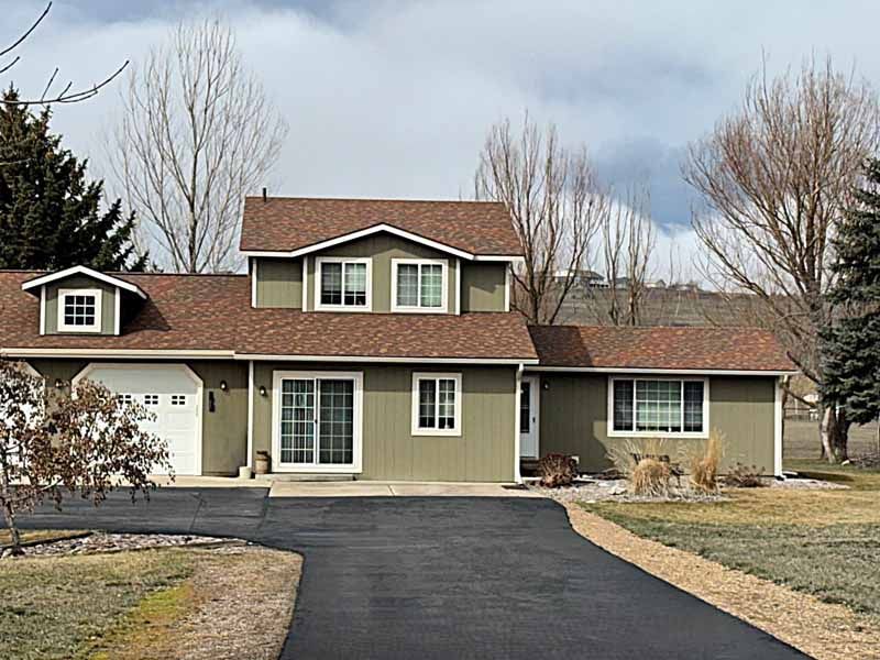 A two-story olive green house with a brown shingled roof, an attached garage, and a paved driveway under a cloudy sky.