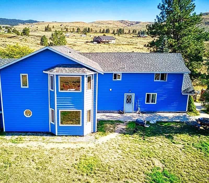 A bright blue two-story house with white trim stands in a dry, grassy landscape with scattered pine trees.