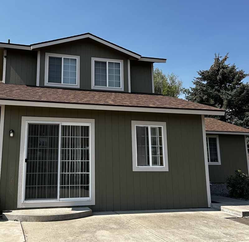 A two-story olive green house with brown shingles, a sliding glass door, and white-trimmed windows under a clear blue sky.
