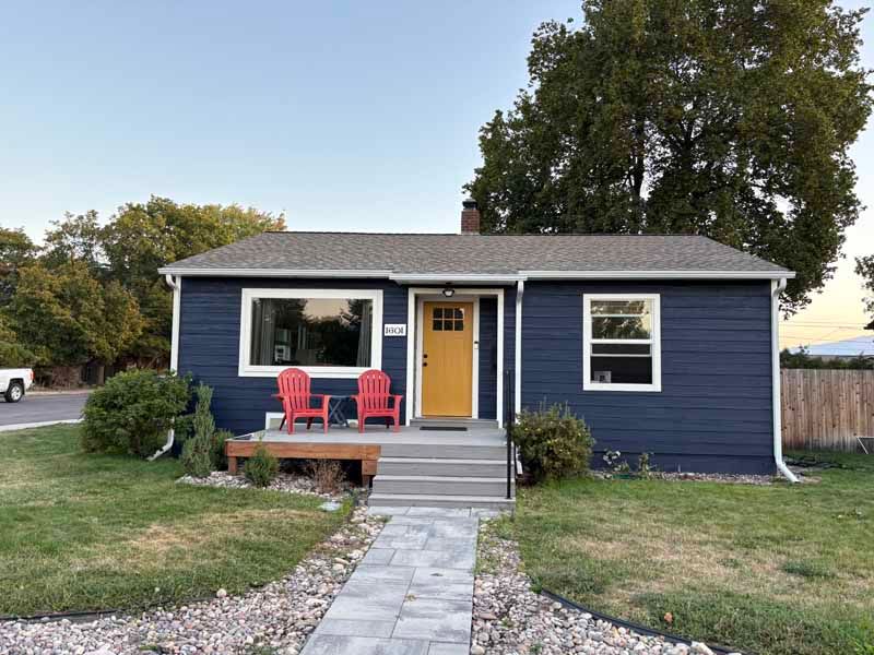 A small, one-story house with dark blue siding, a bright yellow door, and two red chairs on a small front porch.