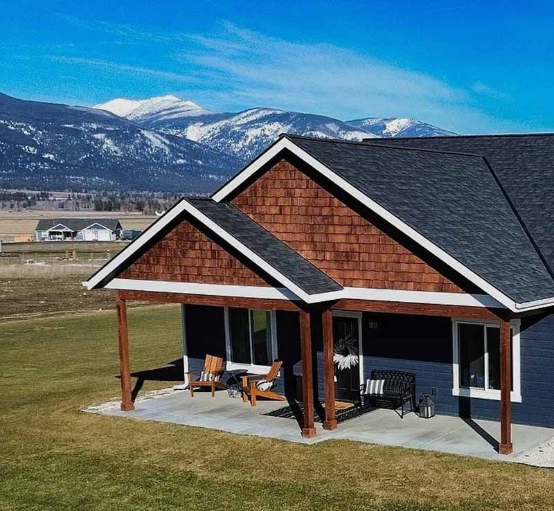 A house with a dark blue lower exterior and brown shingle gable, featuring a concrete porch against snowy mountains.