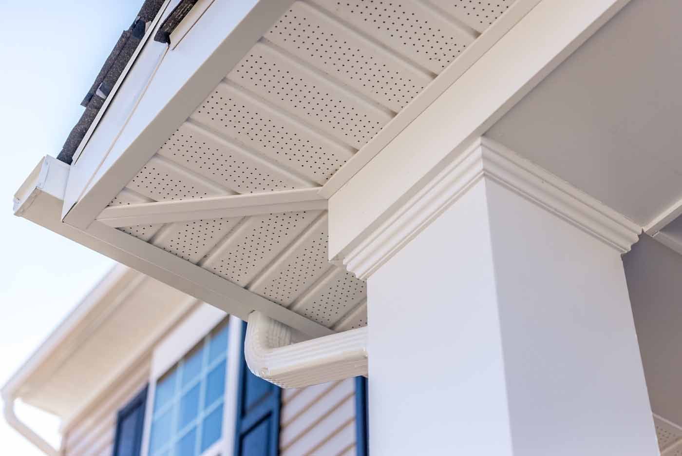 A low-angle view of a white porch column with molding, meeting a white perforated vinyl soffit and a small downspout.