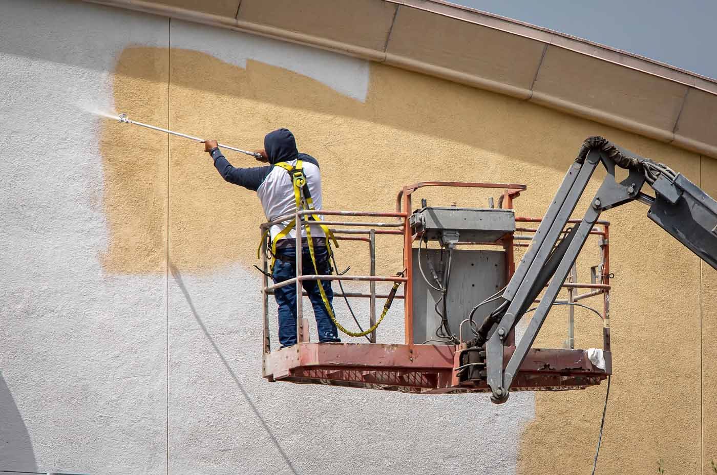 A worker in a safety harness uses a spray gun to paint a tan, textured exterior wall from an aerial lift.