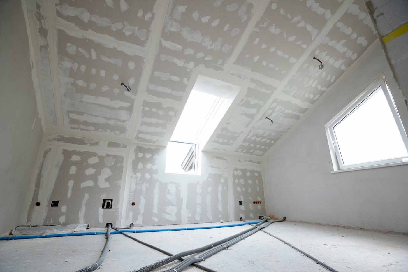 An unfinished attic room featuring grey drywall, exposed wiring, a skylight, and a window against a white concrete floor.