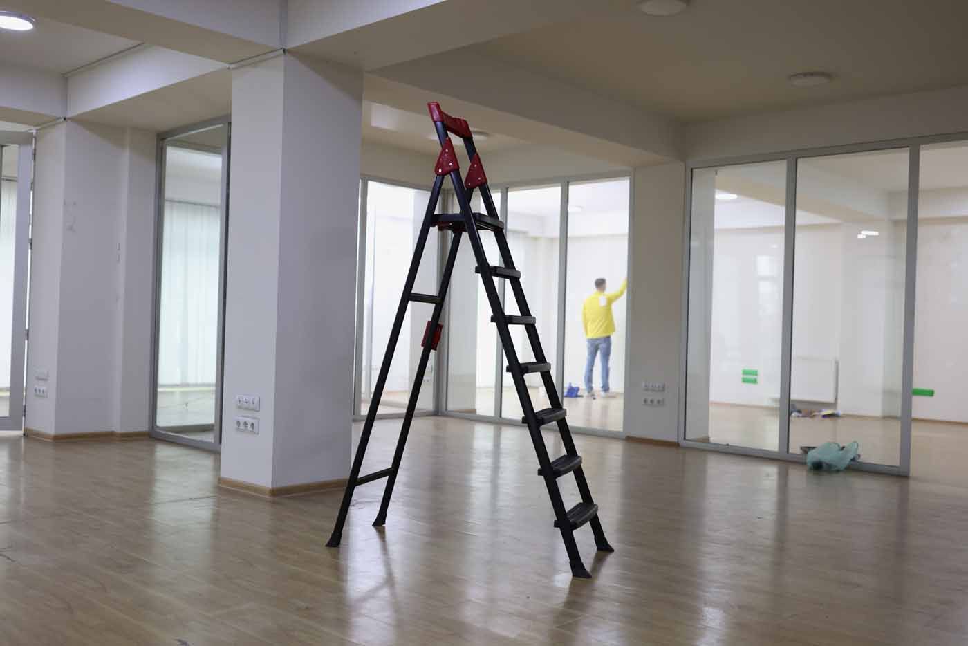 A step ladder stands in the center of an empty office, while a person in a yellow jacket works in the background.