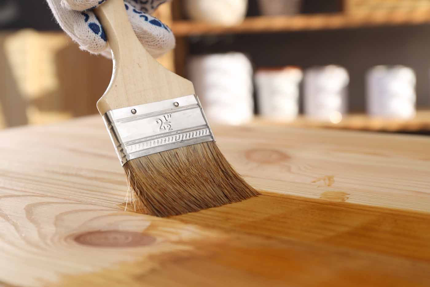A gloved hand uses a brush to apply a warm, amber-colored wood stain to a light-colored wooden surface.