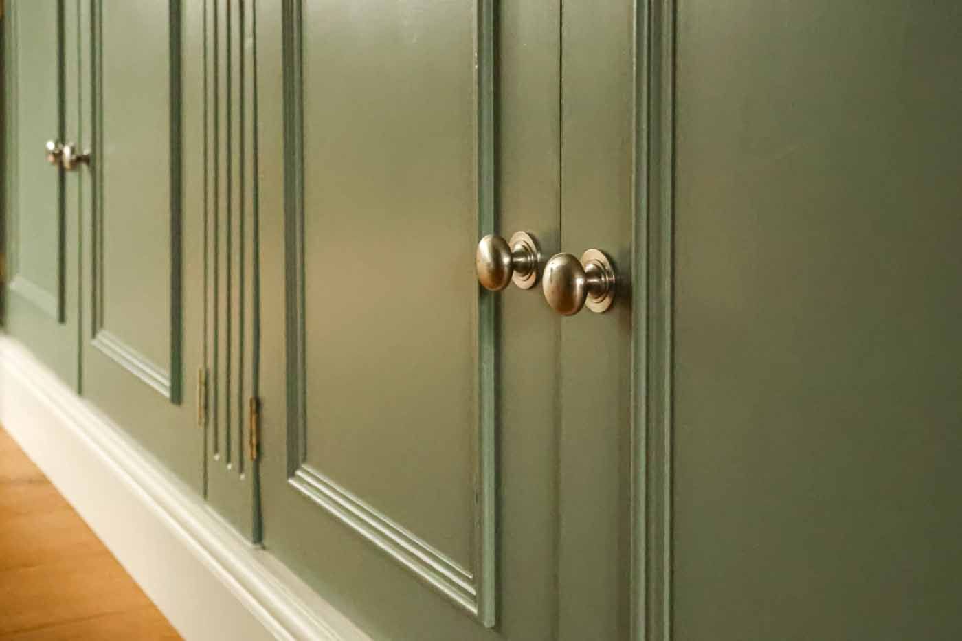 Sage green kitchen cabinetry with silver mushroom knobs and raised panel details on wooden flooring.