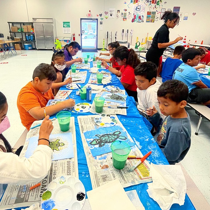 A group of children are sitting at tables in a classroom painting.