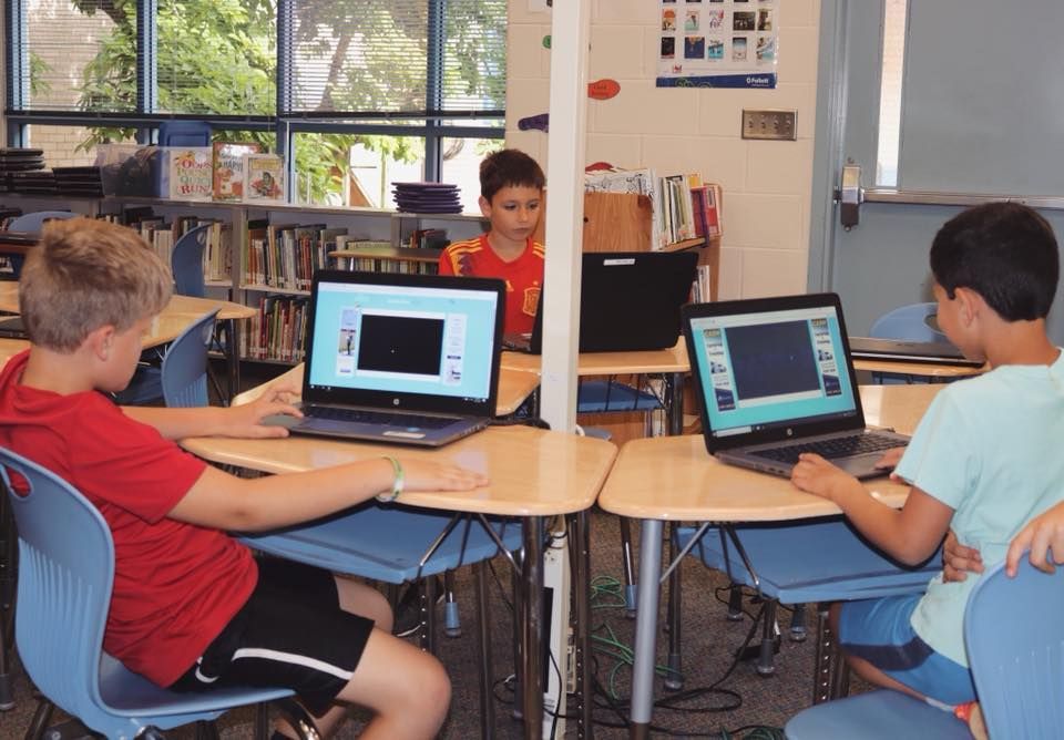 A group of children are sitting at desks using laptops