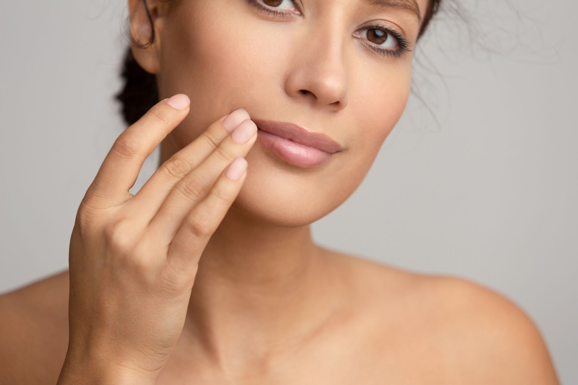 Woman touching her lip with her fingers, gazing towards camera; neutral background.