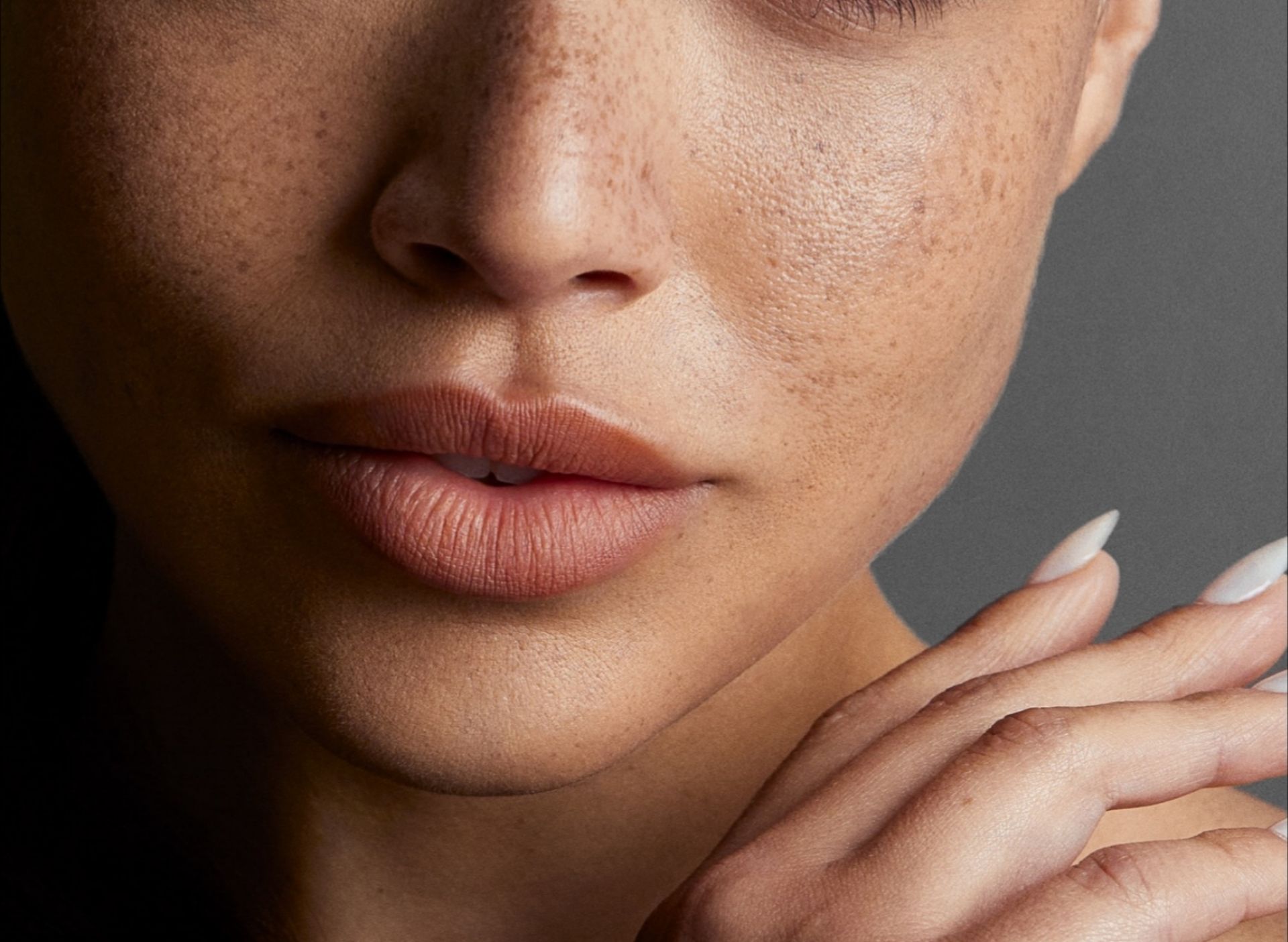 Close-up of a person with freckled skin and natural-toned lipstick, resting a hand with long, light-colored nails on a shoulder.