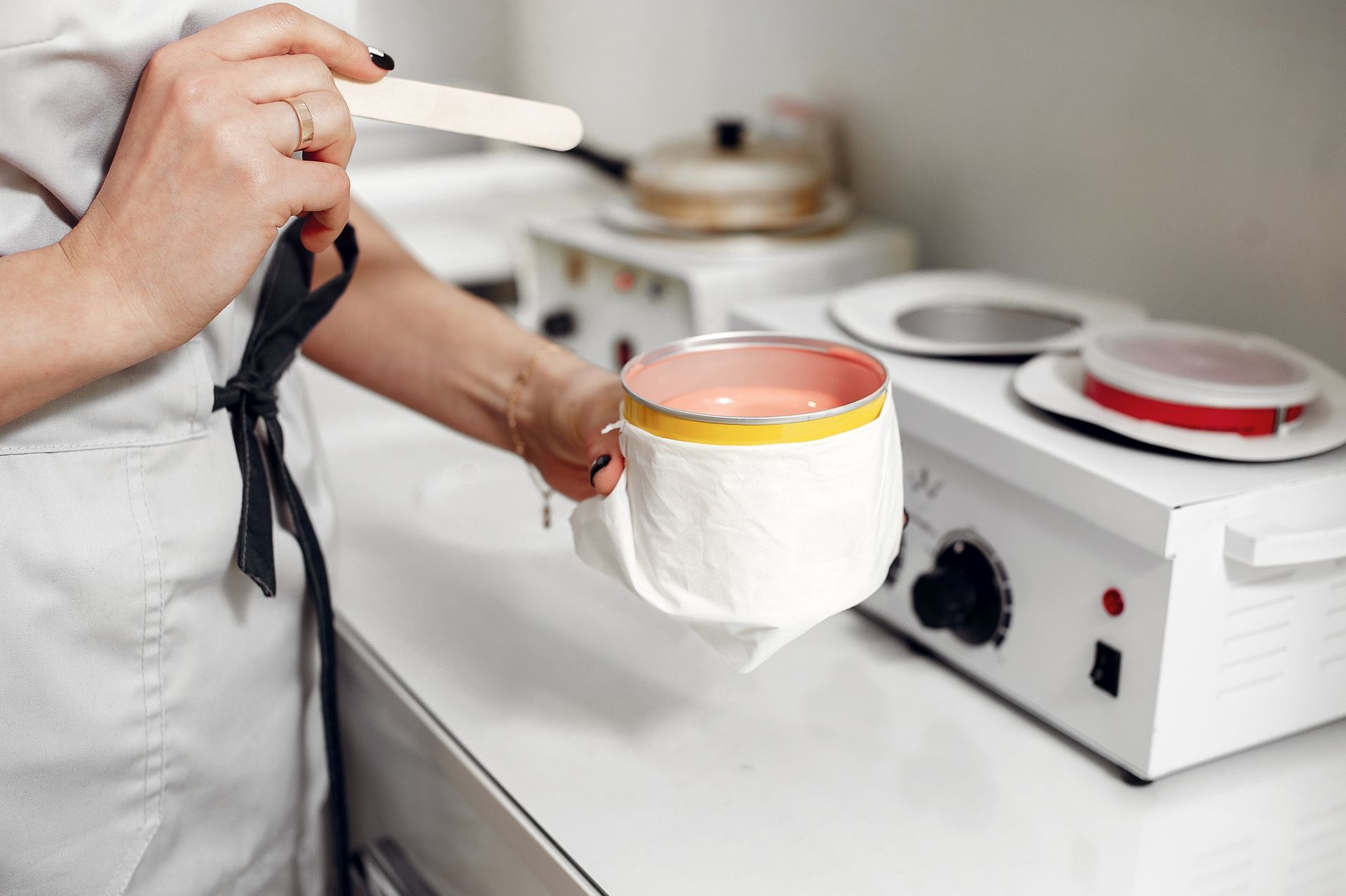 A woman is holding a cup of wax and a spatula.