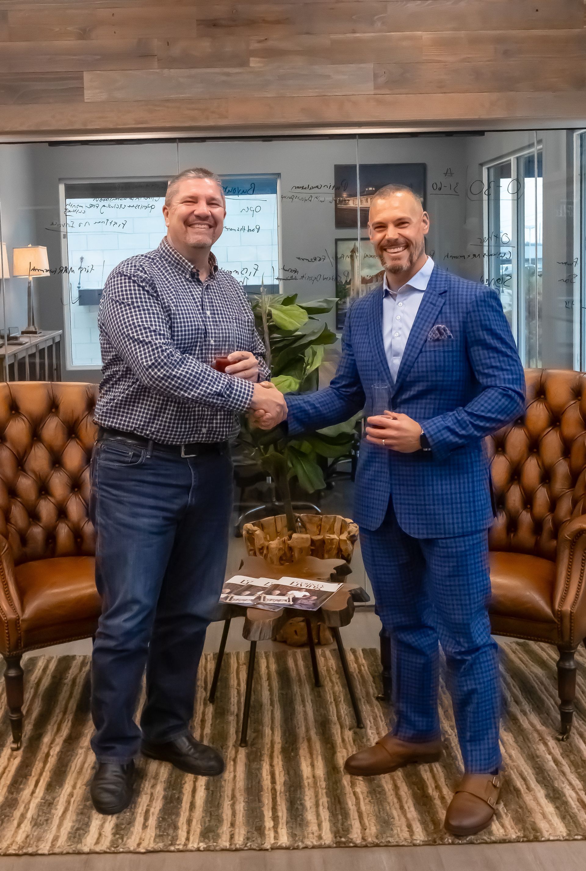 Two men are shaking hands in a living room while holding wine glasses.