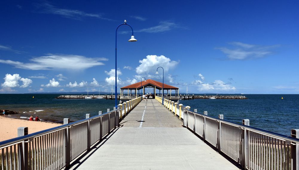 A long wooden pier with metal railings extends over blue water toward a small, roofed pavilion under a sunny, blue sky— Moreton Bay Therapy in Deception Bay, QLD