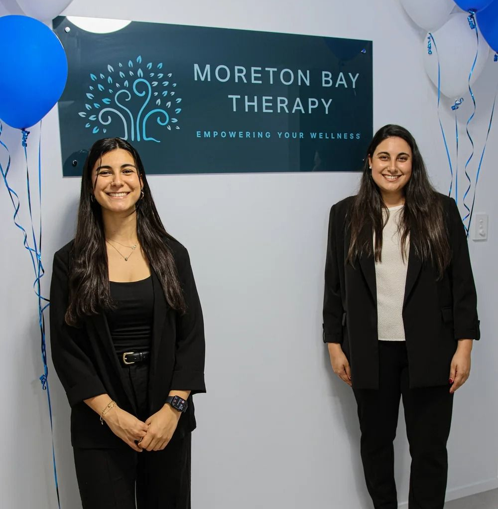Two Smiling People Stand in Front of a Sign — Moreton Bay Therapy in Deception Bay, QLD
