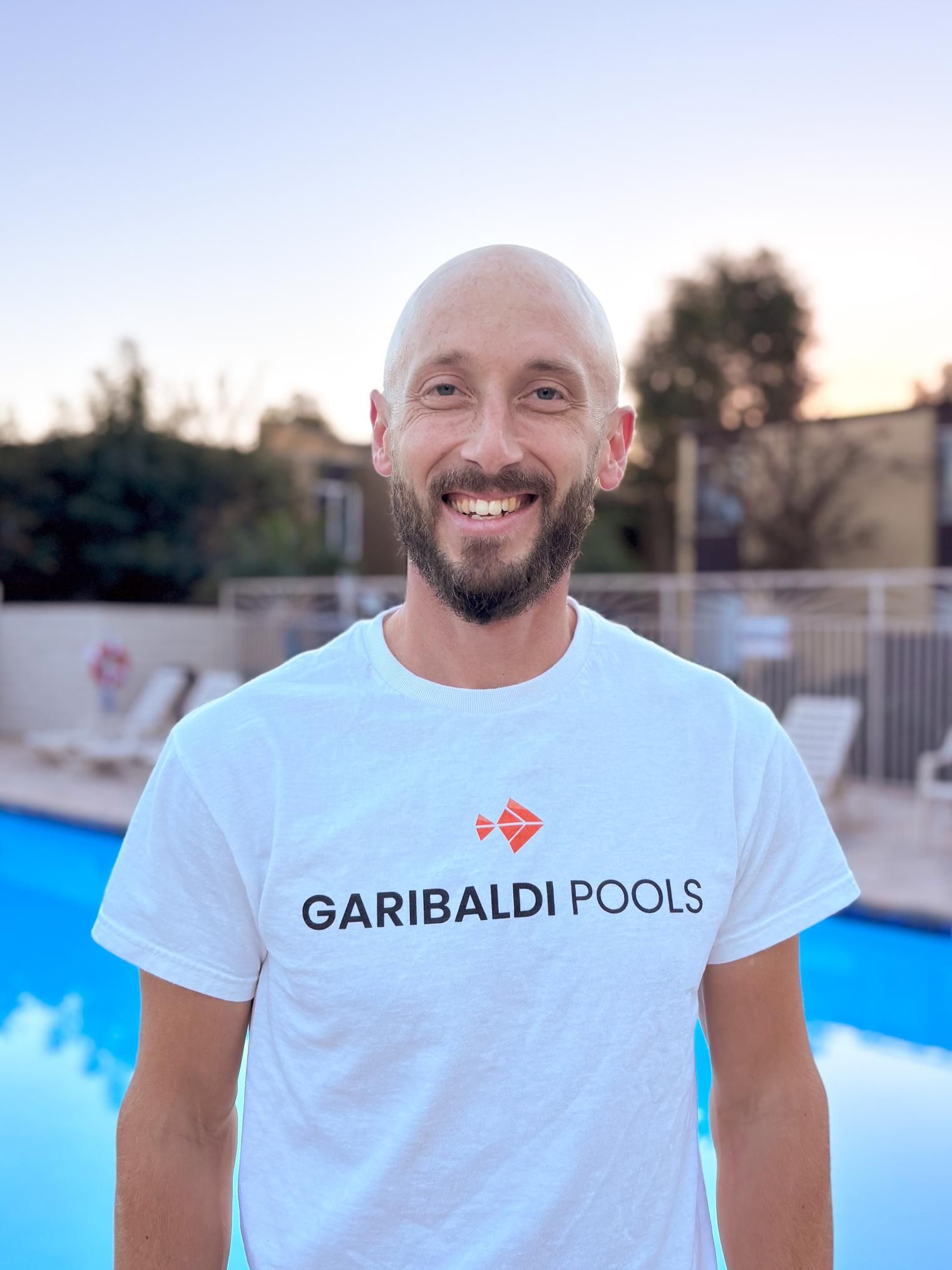 A bald man with a beard is wearing a white t-shirt that says garibaldi pools.