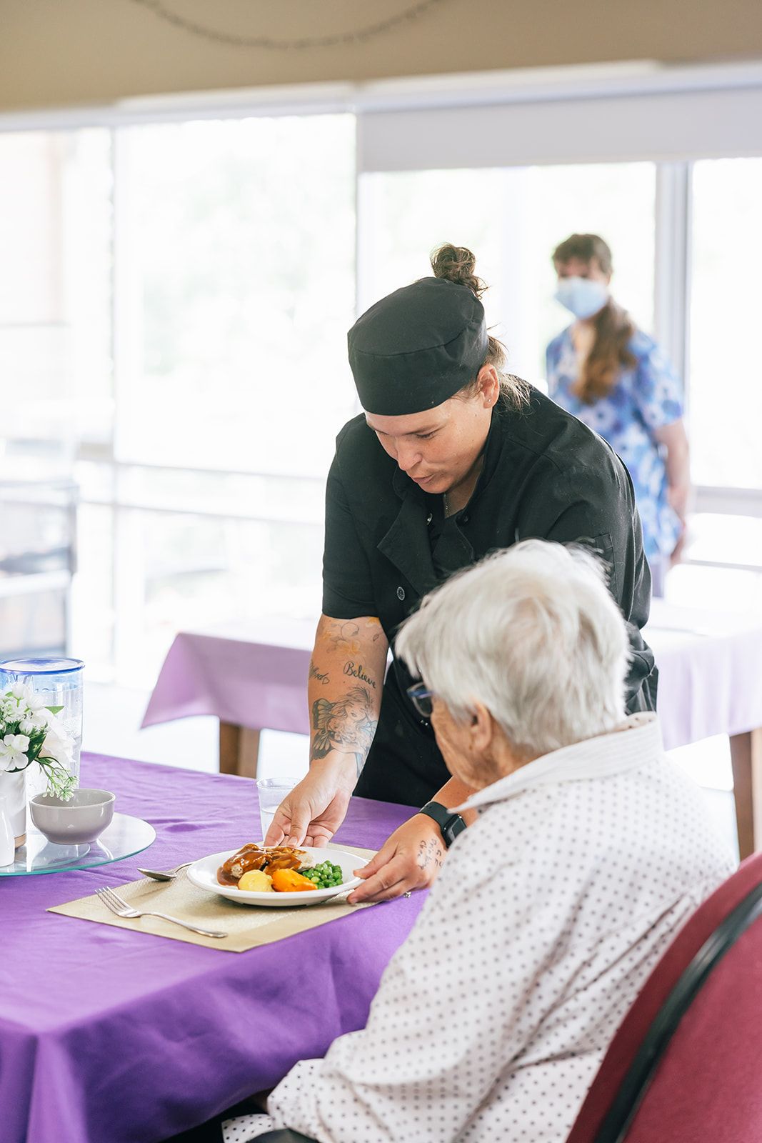 A food service worker in a black uniform places a plate of food on a table before a seated person in a purple-clothed room.