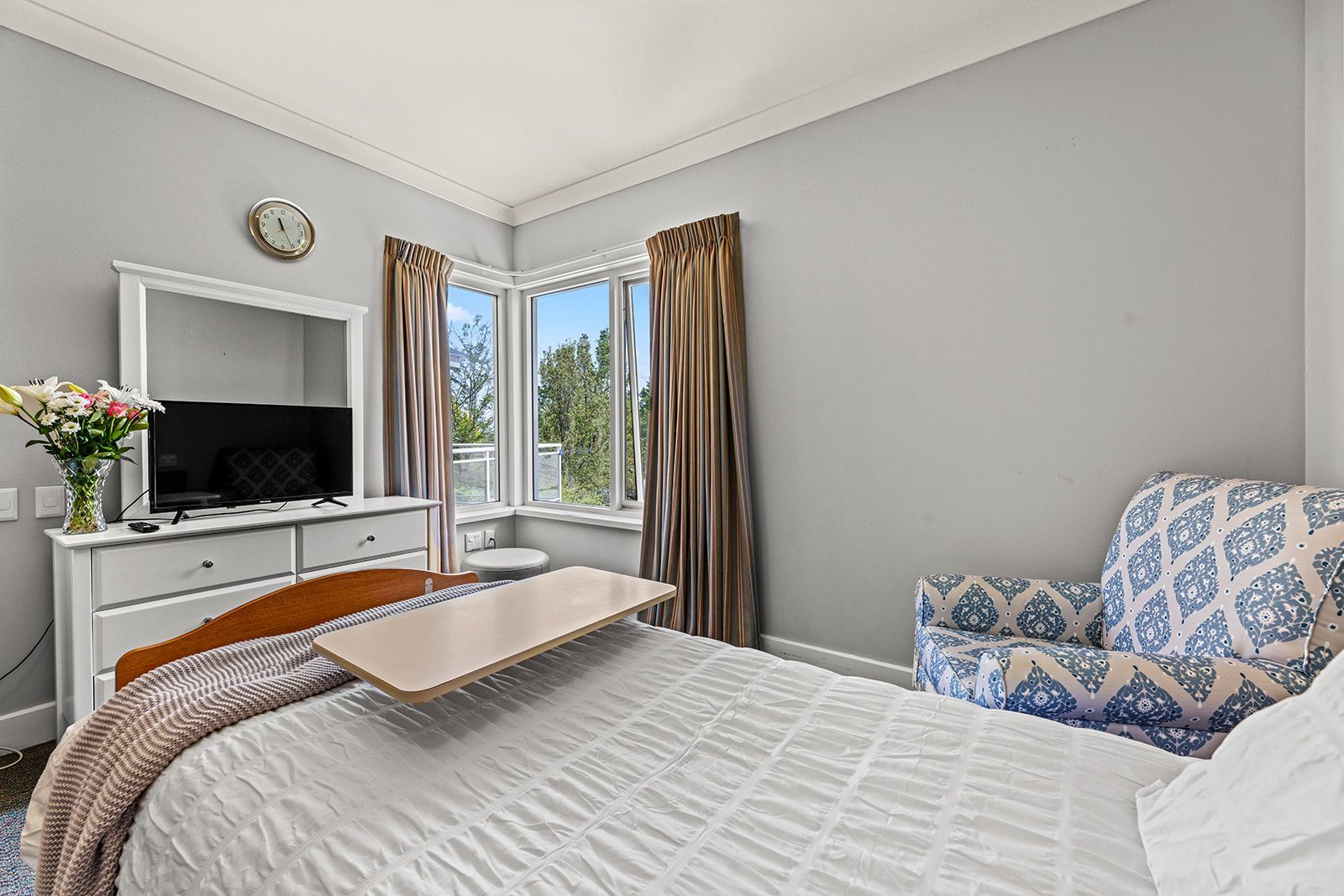 A bedroom with a bed featuring a wooden tray, a white dresser with a television, and a patterned armchair by a window.