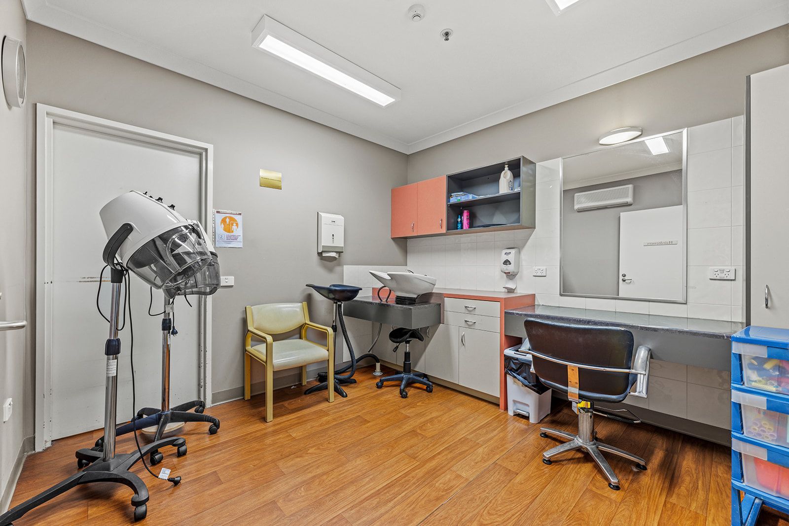 A hair salon room with a floor-standing hair dryer, a wash basin station, a styling chair, and a large mirror.