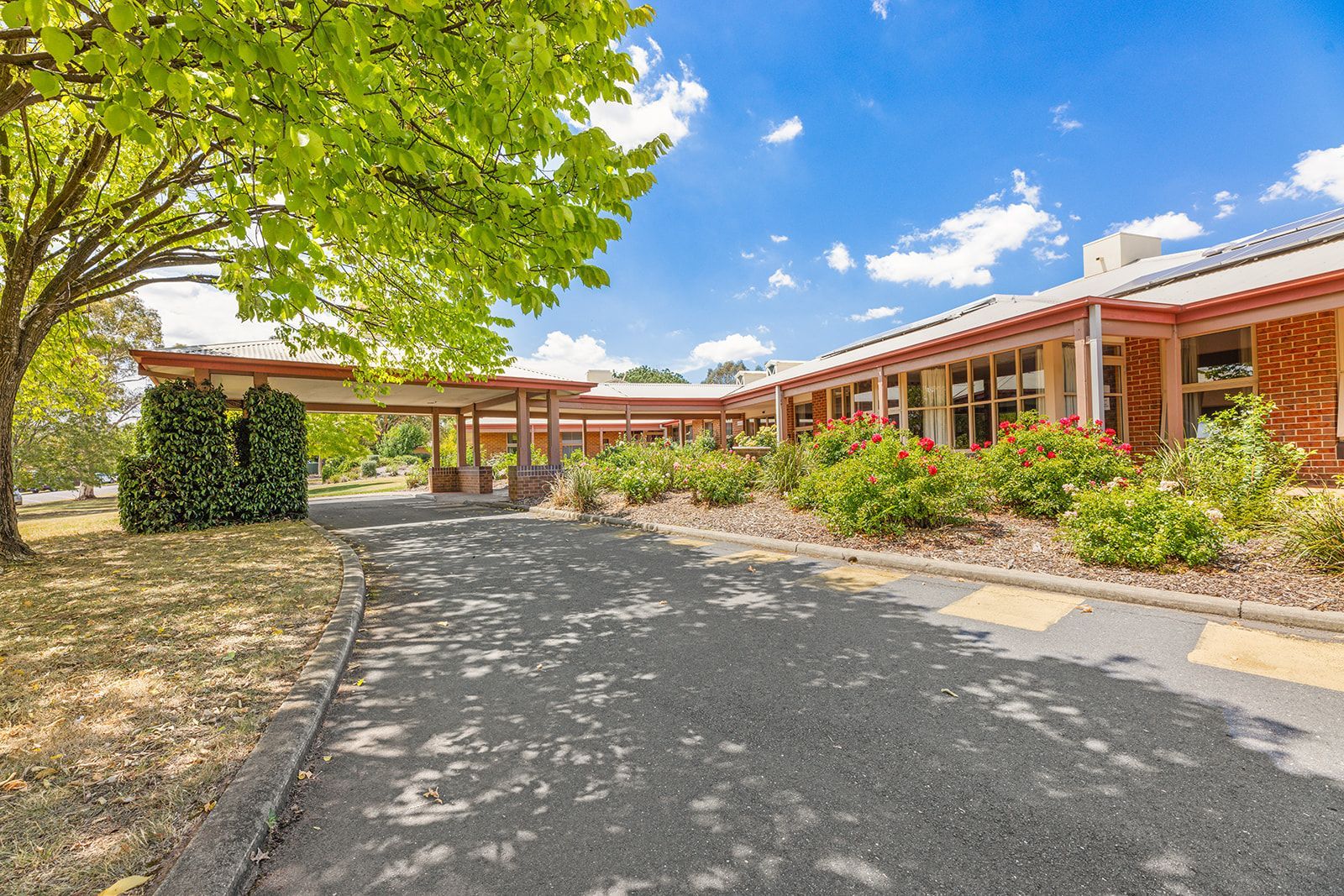 A paved driveway leads to a long, single-story red brick building with a covered entrance under a large leafy tree.