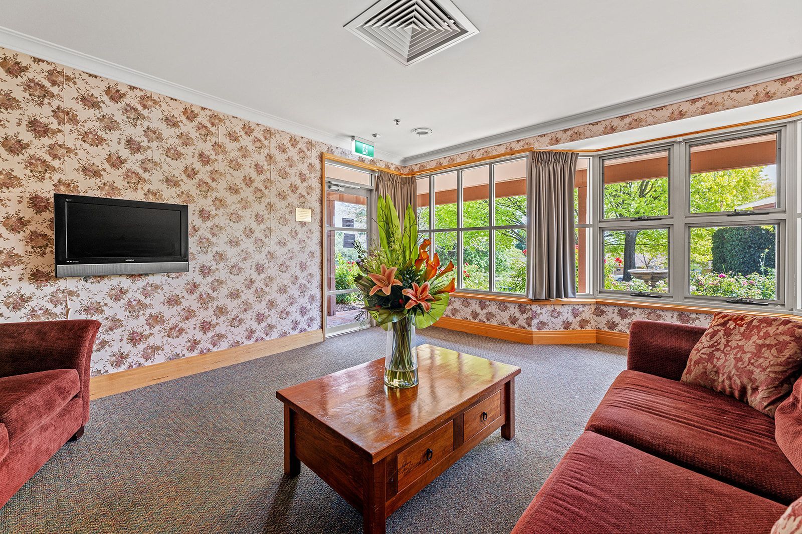 A carpeted lounge featuring patterned wallpaper, two red couches, a central wooden coffee table with flowers, and windows.