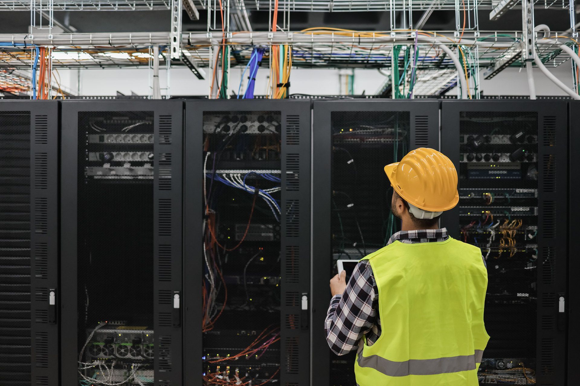 Technician in yellow vest and hardhat inspecting server racks in a data center.