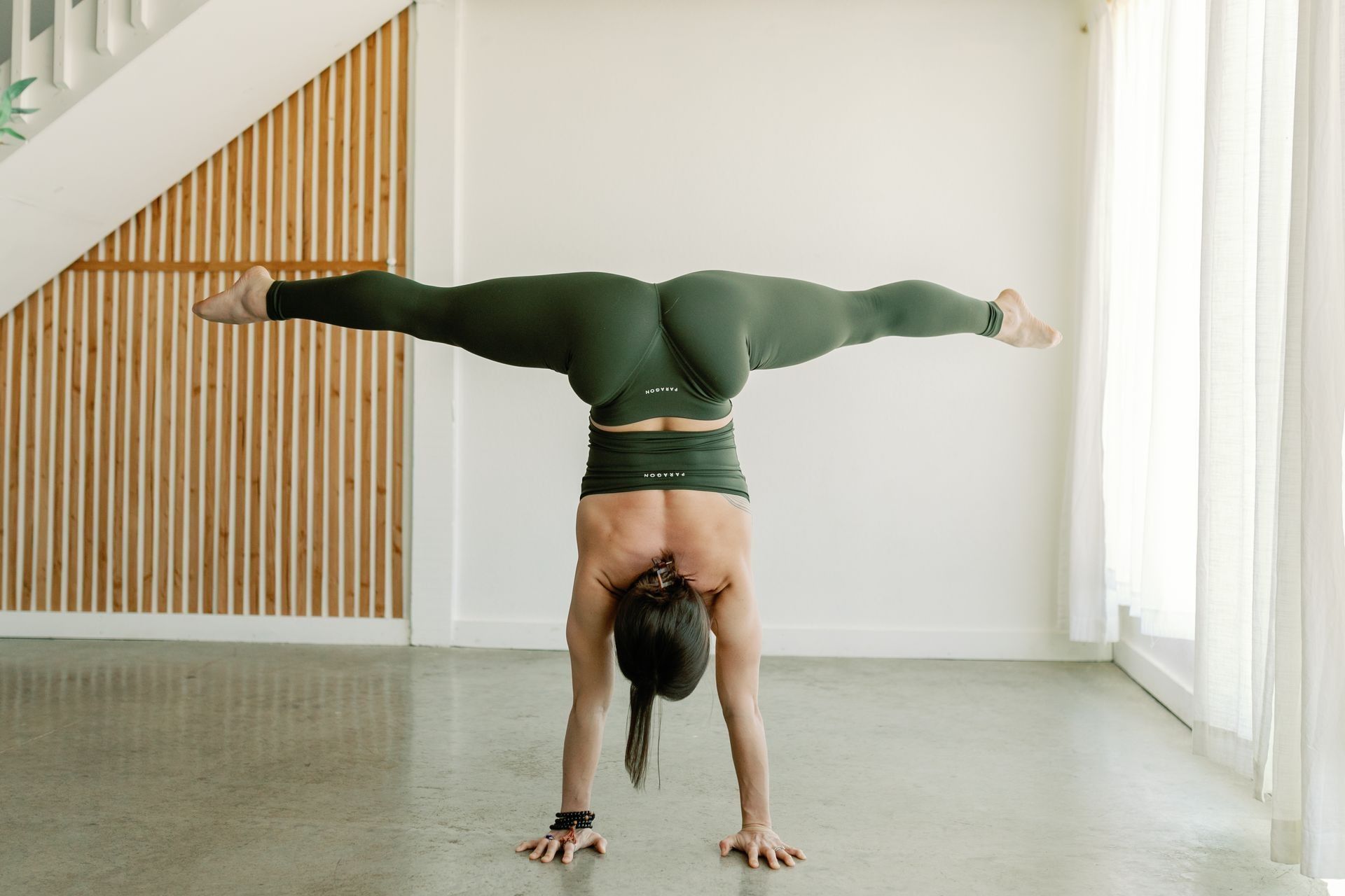 A person in dark green athletic wear performs a handstand with a wide straddle split in a bright, modern studio.