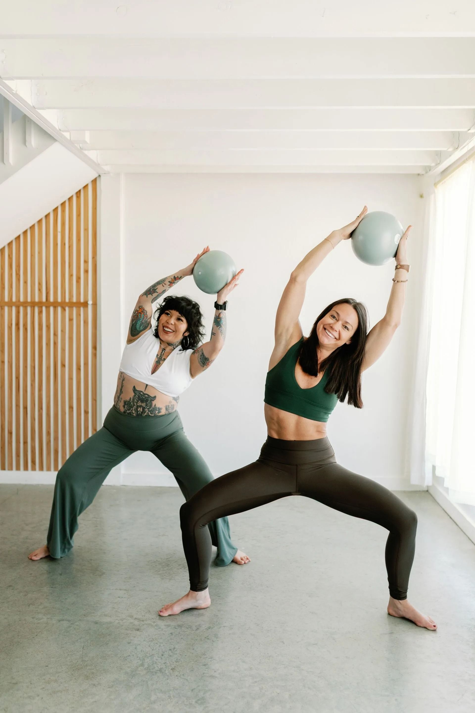 Two people in fitness attire performing side bends while holding small exercise balls in a bright studio.